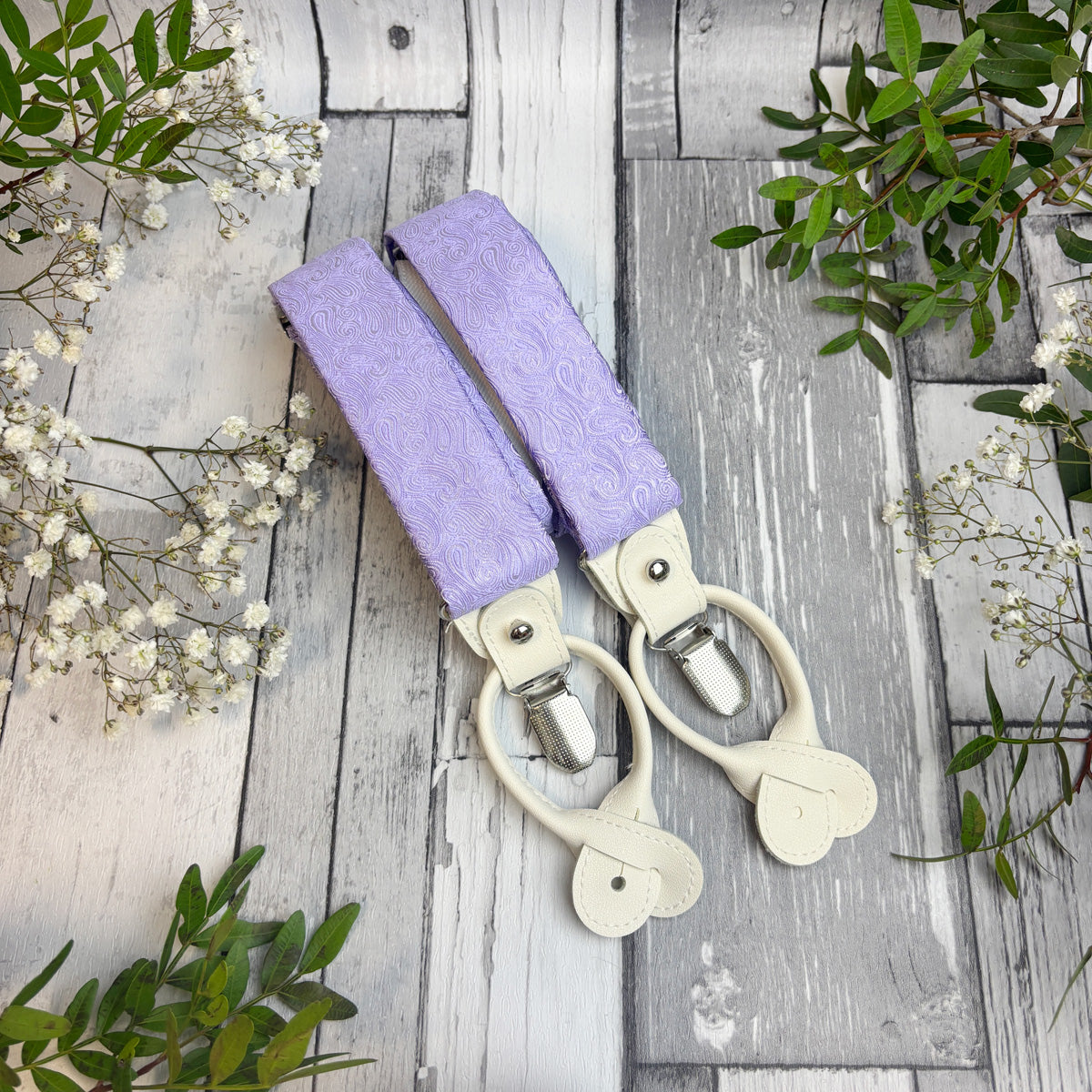 A pair of suspenders with lavender purple patterned fabric straps and cream-colored plastic clips, laid flat on a rustic gray wooden plank background with baby's breath flowers and green leaves.