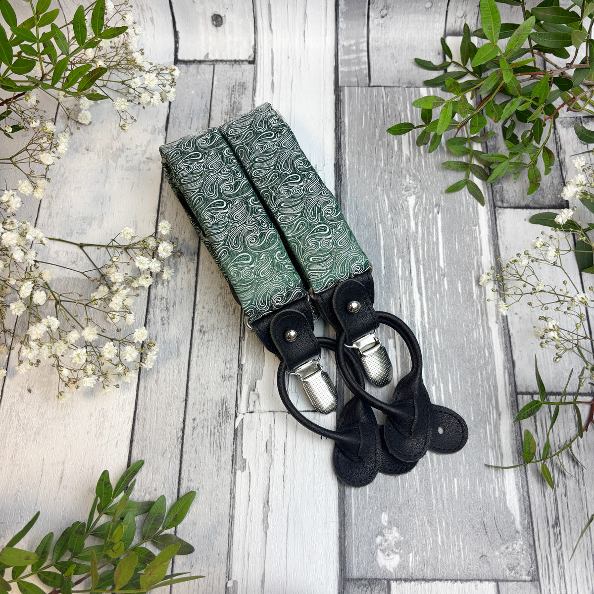 A pair of green patterned fabric suspenders with black leather loops and silver metal clips, laid flat on a rustic white-washed wooden surface with small white flowers and green leaves around the edges.