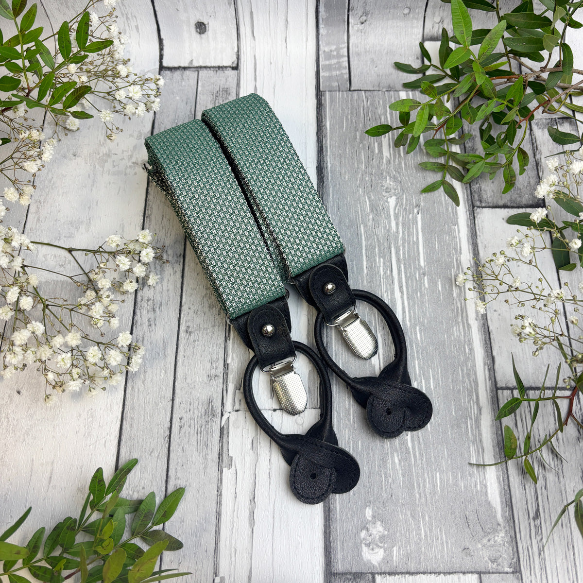A pair of green patterned fabric suspenders with black leather loops and silver metal clips, laid flat on a rustic white-washed wooden surface with green leaves and small white flowers as decorative props.