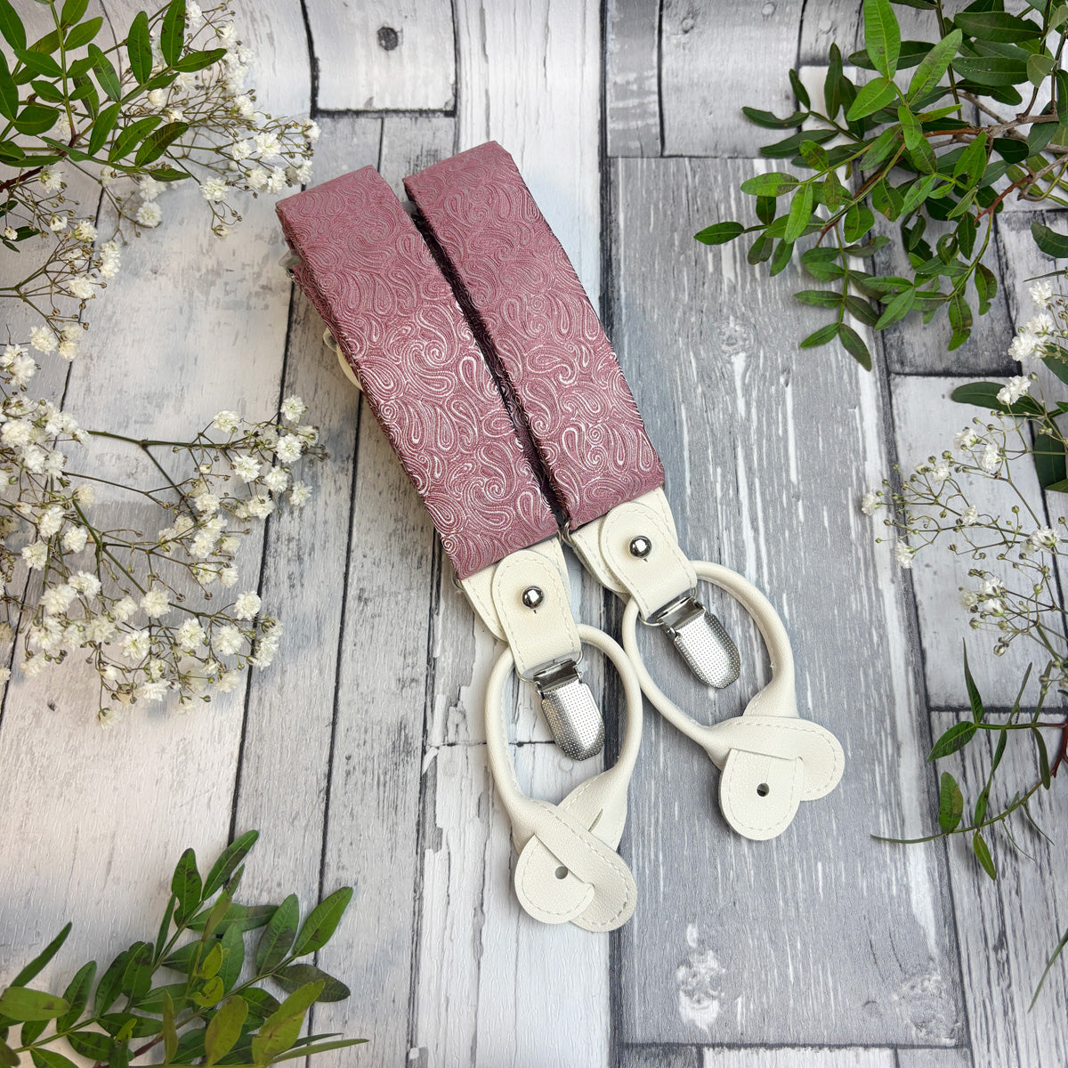 A pair of dusty rose leather end braces with silver clips, arranged on a rustic gray wooden plank background with decorative green leaves and white baby's breath flowers.