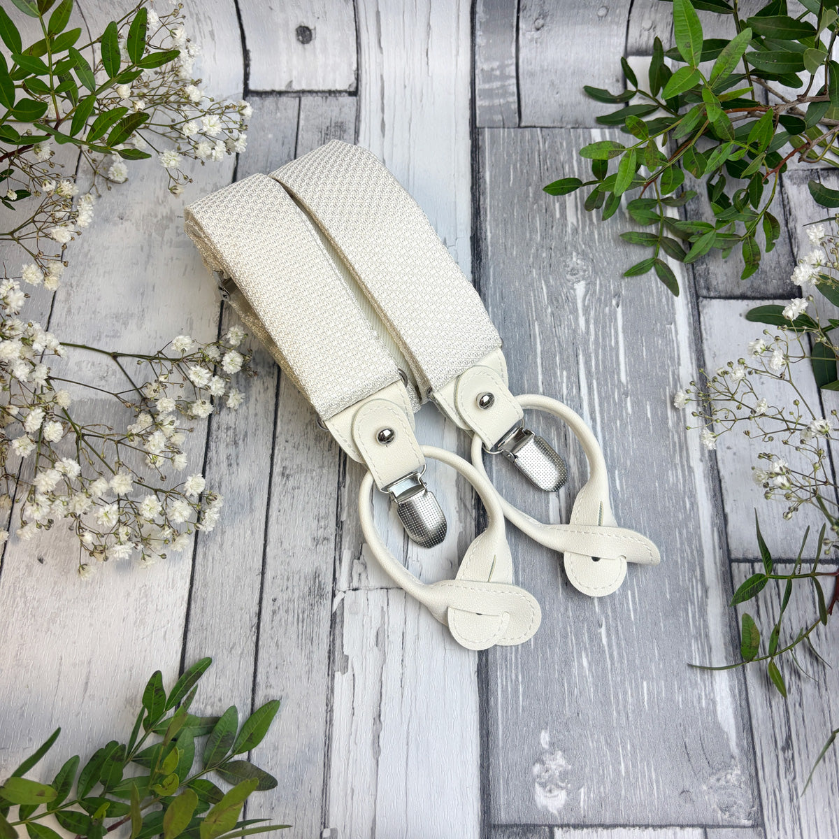 Off-white woven fabric suspenders with silver metal clips, styled on a rustic white-washed wooden background with green leaves and white baby's breath flowers.