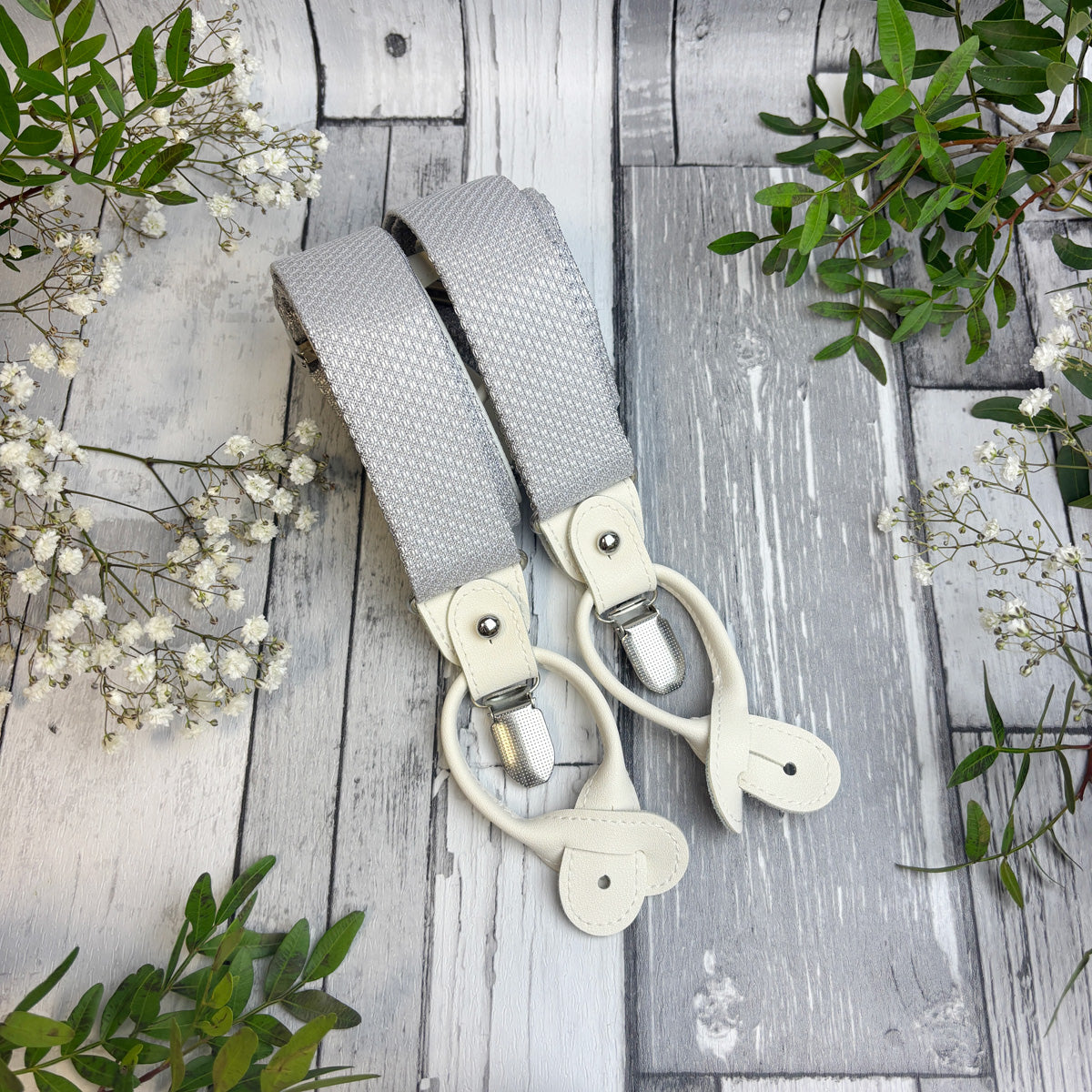 A pair of light gray woven fabric suspenders with cream-colored leather clips, arranged on a rustic gray wooden plank surface decorated with green leaves and white baby's breath flowers.