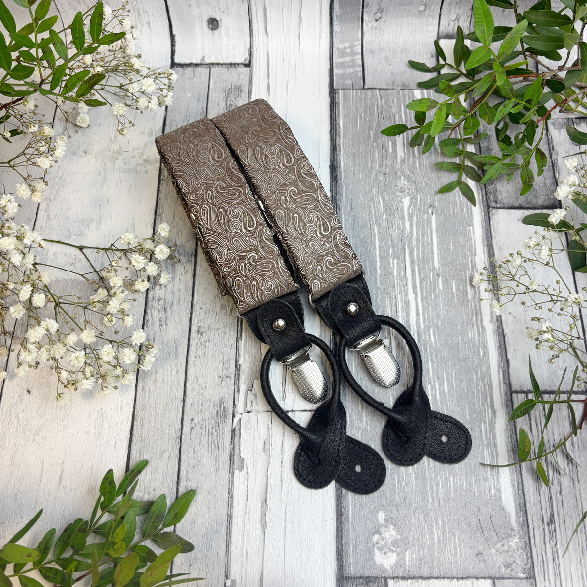 A pair of brown paisley patterned suspenders with black leather clips and silver metal rings, laid flat on a rustic white-washed wooden surface with green leaves and white flowers as decorative props.