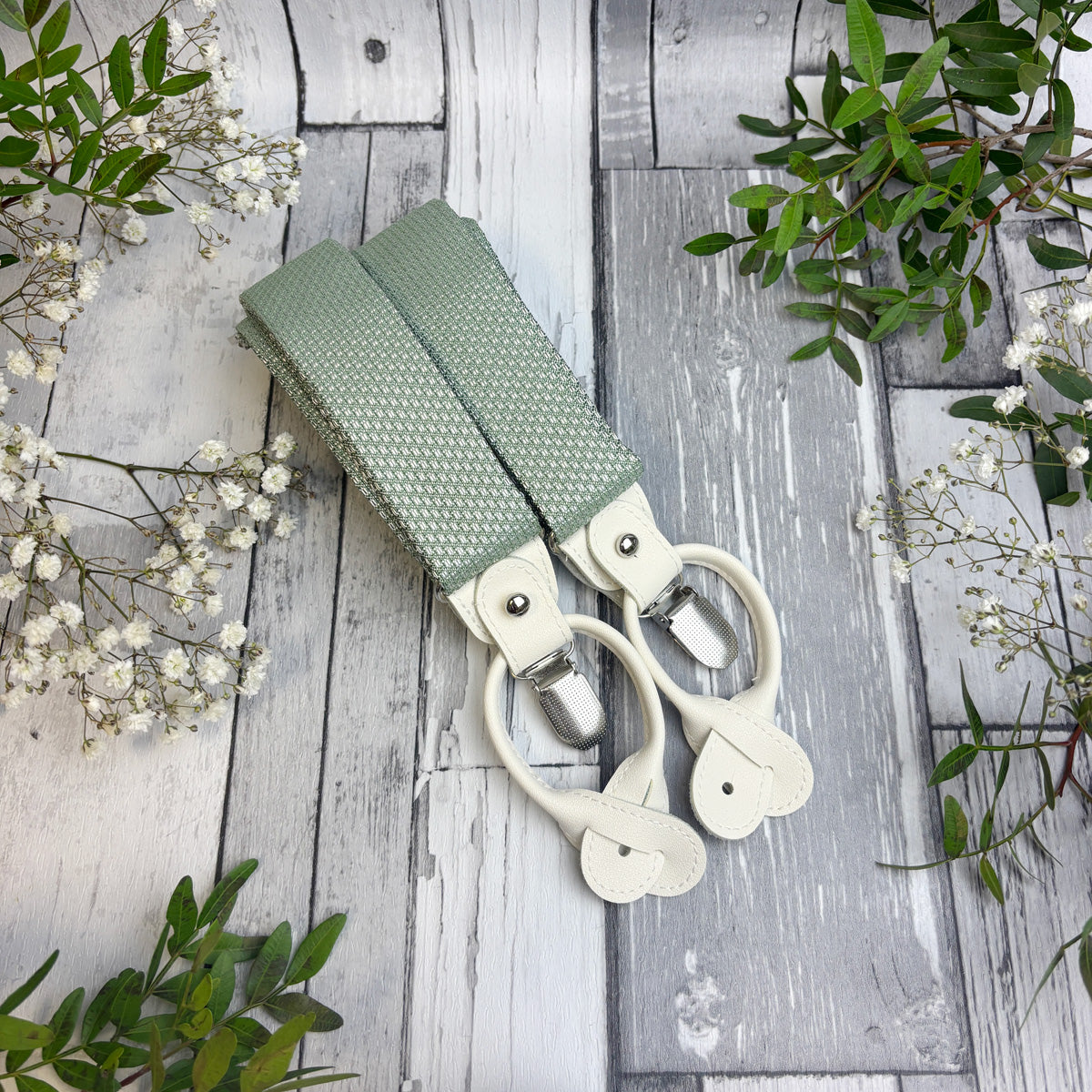 A pair of sage green patterned fabric suspenders with cream-colored plastic clips, laid flat on a rustic gray wooden plank surface decorated with white baby's breath flowers and green leaves.